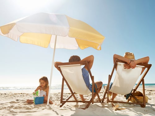 A child plays with a bucket and spade on a sunny beach, while two adults relax on deck chairs under a yellow umbrella, creating a peaceful scene.