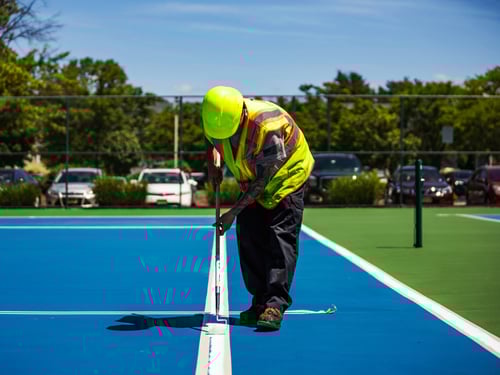 paving contractor adding striping and boundary lines on a new, blue outdoor tennis court installation