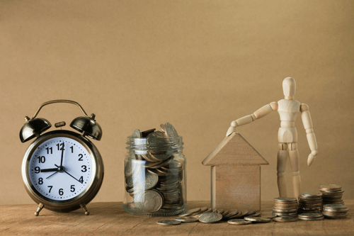 A vintage bell alarm clock, glass jar of coins, house-shaped wooden block, and wooden mannequin arranged side by side.