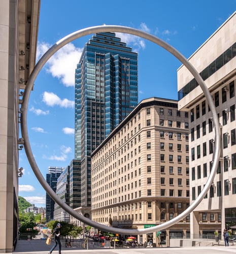 View of a city with a large circular metal sculpture framing skyscrapers under a bright blue sky. A person walks, conveying a lively urban vibe.