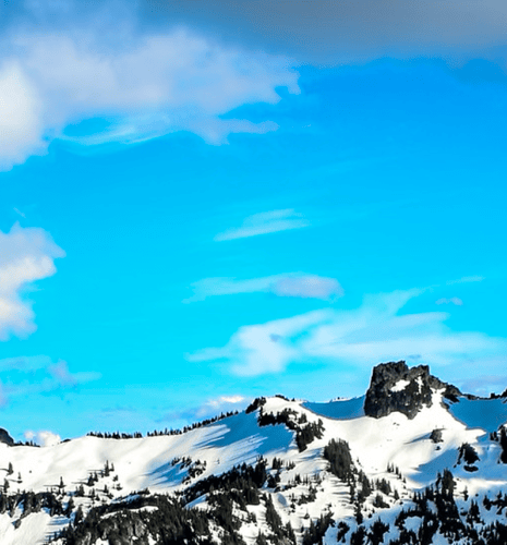 Snow-covered mountain landscape under a bright blue sky with scattered clouds. Sunlight casts shadows, creating a peaceful, serene atmosphere.