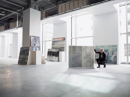 A person in a bright, spacious art gallery examines a large abstract painting. Sunlight streams through tall windows, illuminating several artworks.