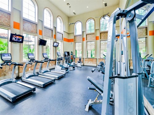 A modern style fitness center with tall windows and  fully equipped with fitness equipment at Mission at La Villita Apartments, Irving, TX