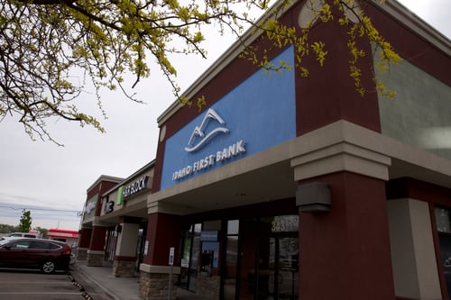 The exterior of a commercial building featuring an "Idaho First Bank" branch. The bank has a blue section with its logo and name, and the building features red and light green/grey sections with stone accents at the base.