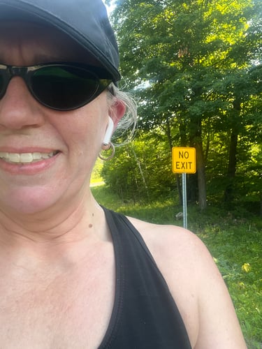 Selfie of Colleen on a Sunny day,  A yellow "NO EXIT" road sign behind her.