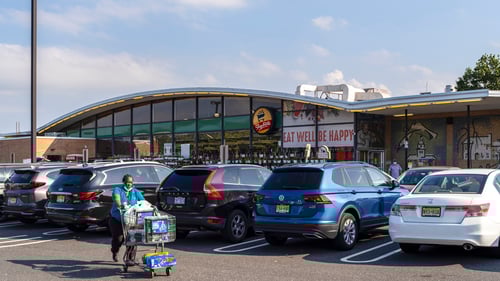 Person pushing shopping cart through full parking lot in front of Shop Rite