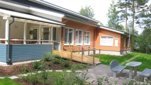Ulkokuva Multatiestä. Rakennus on oranssihtavaa puuta, paitsi terassin osalta seinät ovat valkoiset ja kaiteet siniset. Terassilla ja pihalla on pöytäryhmät. Pihalla on istutuksia ja vehreä nurmikko.