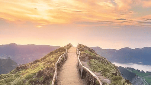 A winding path with a wooden railing leads towards a sunset over rolling hills and a distant body of water. Soft pastel skies above.