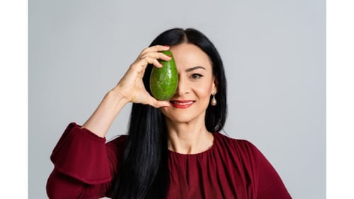 Anamaria Hager, Expertin für Chronobiologisches Ernährungstraining (CbE), hält lachend eine grüne Avocado vor ihr rechtes Auge. Sie trägt ein weinrotes Oberteil mit gerafftem Ausschnitt, ihre langen dunklen Haare fallen offen über die Schulter. Das Bild vermittelt Humor, Nahbarkeit und Freude an natürlicher, gesunder Ernährung.