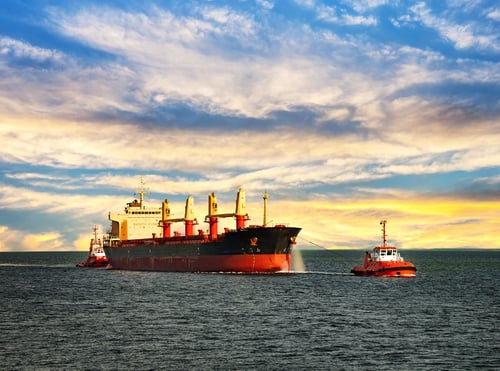 Large cargo ship with red and black hull being assisted by tugboat at sea at sunset.