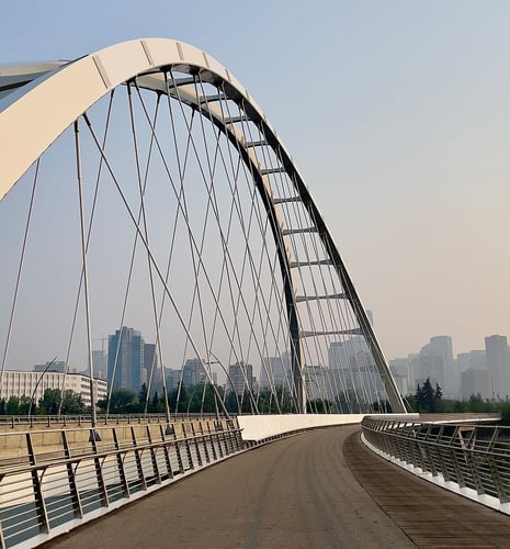 A modern, arched pedestrian bridge with cable supports leads into a distant city skyline. The scene is calm, with a clear sky and scattered buildings.