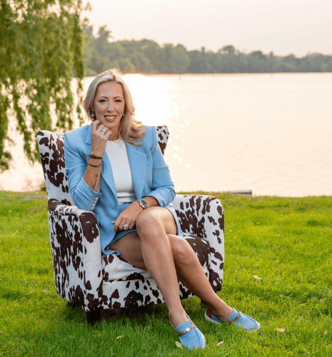 Caitlin Burton in a blue blazer seated on a cow-print chair by a serene lake at sunset. She smiles warmly, exuding a relaxed and confident vibe.