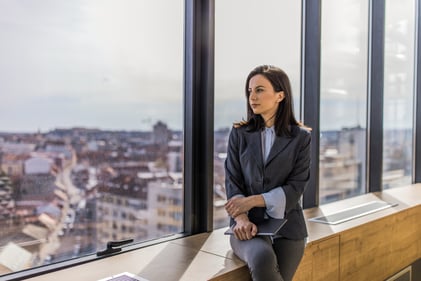 professionally dressed woman staring pensively out of a skyscraper's window with city in background