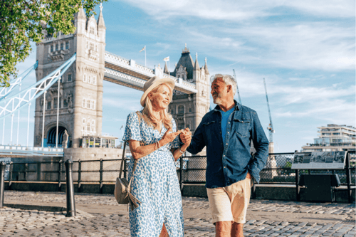 Elderly couple walks hand in hand near London's Tower Bridge on a sunny day, smiling and enjoying each other's company in a joyful atmosphere.
