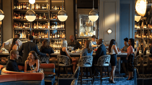 Elegant bar with guests socializing, bartenders preparing drinks, plush seating and shelves stocked with liquor bottles under warm lighting