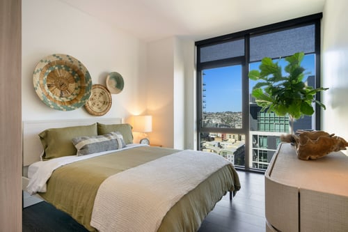 Bright bedroom with green bedding, basket-style wall art, and floor-to-ceiling windows showcasing city views.