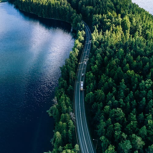 Aerial view of a winding road cutting through dense green forest beside a tranquil blue lake. Cars travel along the road, conveying a sense of peaceful journey.