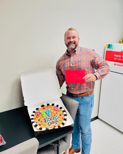 Agent Hank holding a red card and next to him is a black table with a cookie cake on it
