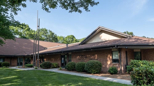 The Church of Jesus Christ of Latter-day Saints meetinghouse in Dalton, Georgia. The viewer is presented with a dark brick building and brown-shingled roof. The building has two sets of glass door entryways, with the name of the church mounted directly on the meetinghouse brick. A white walkway leads from the parking lot to the different sets of doors.Three ornamental metal spires are installed in front of the building. The building is situated on a corner lot.