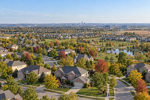 Elevated view of Omaha metro neighborhoods with tree-lined streets, autumn foliage, a community pond, and a distant city skyline.