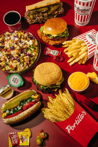 A colorful overhead spread of Portillo’s menu items on a bright red background, featuring crinkle cut fries, a Chicago style hot dog, burgers, an Italian beef sandwich, a chopped salad, cheese sauce, drinks, and dipping sauces arranged around the table.