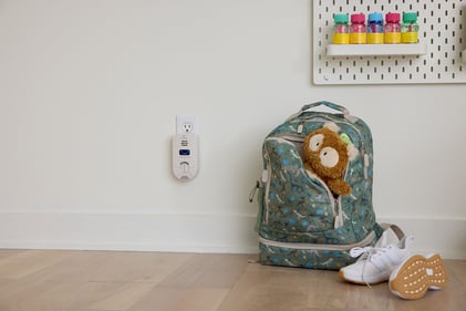 Child's backpack and shoes on the floor next to a wall outlet with a carbon monoxide alarm.