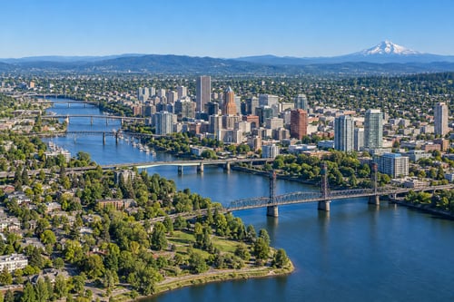 Aerial view of Portland’s skyline, Willamette River bridges, and leafy neighborhoods, with Mount Hood in the distance beneath a clear blue sky.