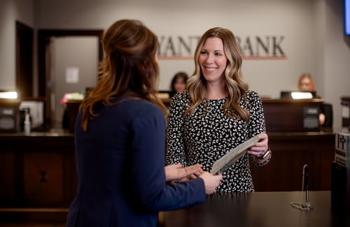 A smiling Bryant Bank employee in a leopard-print blouse assists a customer at a polished wood teller desk. The customer, viewed from the back, is wearing a navy blazer and holding documents during the interaction.