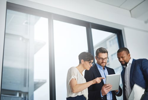 Three professionals collaborate, discussing something on a tablet in a bright, modern office. They appear focused and engaged in teamwork.