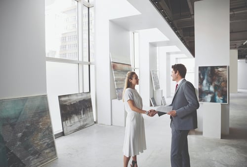 A woman in a white dress and a man in a gray suit shake hands in a bright art gallery with abstract paintings, exuding professionalism and collaboration.