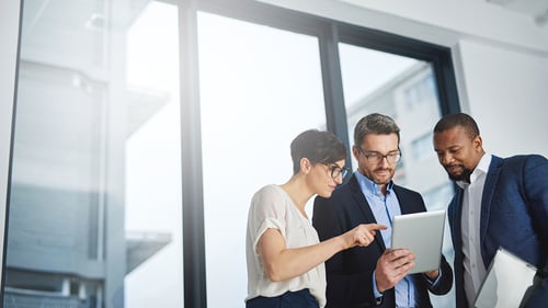 Three professionals collaborate, discussing something on a tablet in a bright, modern office. They appear focused and engaged in teamwork.