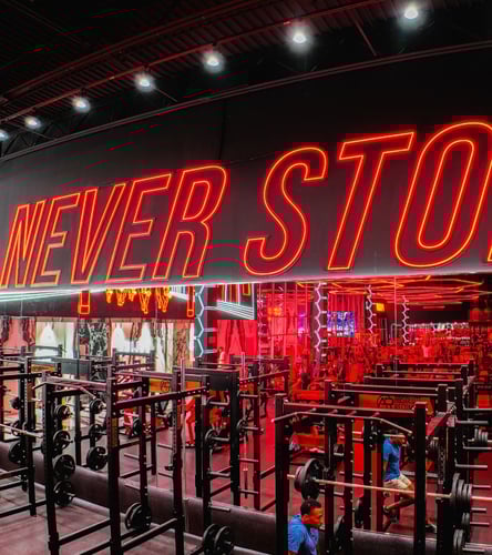 Bold neon red “Never Stop” sign glowing above a vibrant gym floor filled with weight racks, mirrors, and strength training equipment.