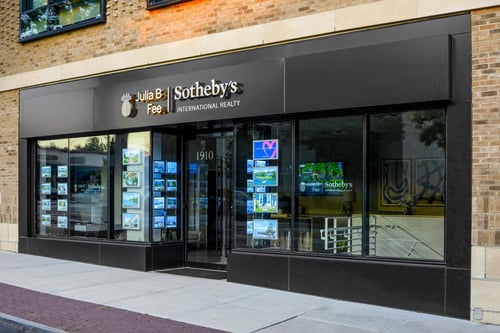 Modern brick building with dark gray storefront and large windows housing Julia B. Fee Sotheby's International Realty in Larchmont, New York, featuring property listing displays.