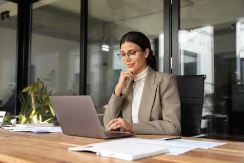 A woman in a beige blazer and glasses sits at a wooden desk, focused on her laptop. Papers are scattered around, and there are plants in the background.