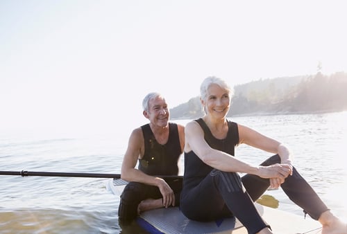 A smiling older couple in wetsuits sit on paddleboards on a calm lake, bathed in sunlight. The scene conveys tranquility and joy.