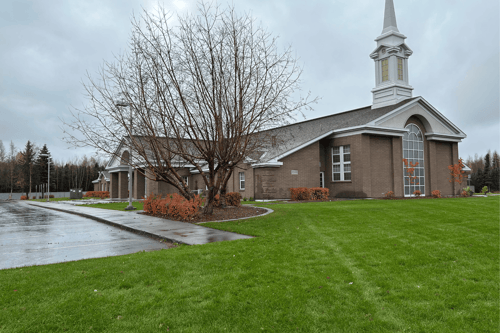 Wide view of a brick church building with a tall white steeple, large arched front window, and a bright green lawn. A leafless tree and orange shrubs sit in the foreground, with wet pavement visible under an overcast sky.
