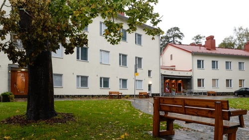 Julkisivukuva kaksikerroksisesta rakennuksesta. Rakennuksen edustalla viherkasveja istutuslaatikoissa, jalankulkukäytävä harmaita laattoja. Talon edustalla myös parkkipaikka, puu ja puisia penkkejä.