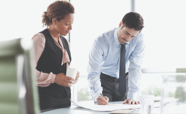 Two professionals in an office setting review a document. One, a woman in a vest holds a mug, while the other, a man in a tie, writes notes. Bright and focused atmosphere.