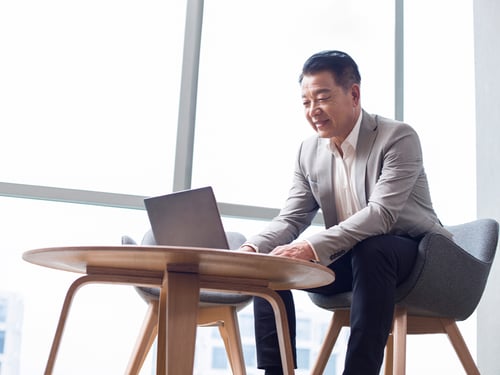 A man in a gray suit sits on a gray chair, smiling and using a laptop on a wooden table. Bright, modern office with large windows in the background.