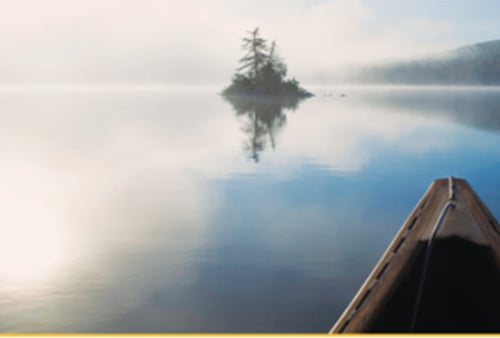A single canoe glides toward a small, tree-covered island on a calm, misty lake. The serene scene reflects tranquility and solitude.