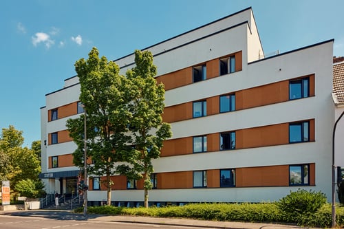 Moderne, mehrstöckige Gebäudefassade mit weißer und ockerfarbener Gestaltung, großen rechteckigen Fenstern und einem Eingangsbereich mit Treppe; davor stehen mehrere grüne Bäume an einer Straße unter blauem Himmel.
