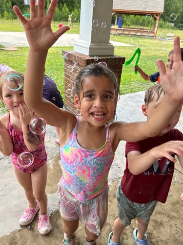 Kids running and playing with floating bubbles in a sunny outdoor space, smiling and laughing.