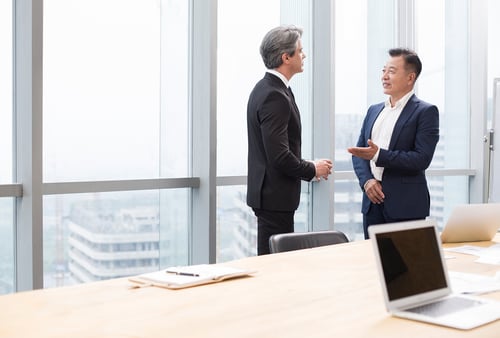 Two businessmen in suits engage in conversation in a bright office with large windows overlooking a cityscape. A laptop rests on a wooden table.