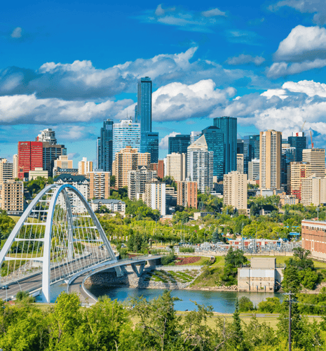 A vibrant city skyline under a bright blue sky with fluffy clouds. A white arch bridge spans a river in the foreground, surrounded by lush greenery.
