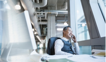 A businessman in a suit smiles while talking on his phone in a modern office. He's seated by large windows, with sunlight streaming in. The scene conveys a professional and positive atmosphere.