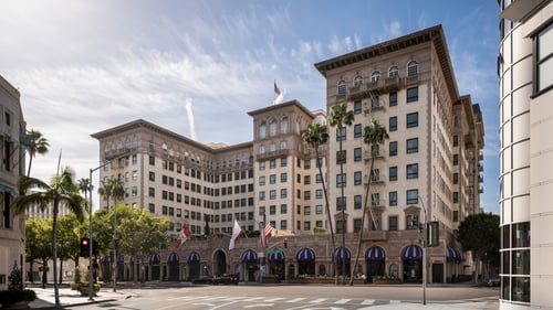 A grand historic hotel with beige brick walls, palm trees, and blue-striped awnings lines a sunny street corner, showcasing elegant architecture and multiple flags.