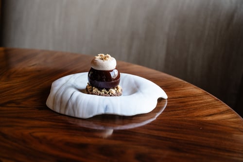 A beautifully crafted dessert featuring a glossy chocolate dome topped with cream and garnished with crumbled nuts, presented on a unique white ceramic dish.