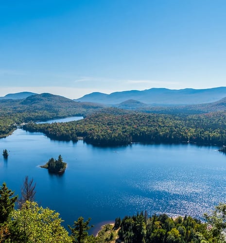Scenic view of a serene blue lake surrounded by lush green forests and distant rolling hills under a clear blue sky, conveying tranquility.