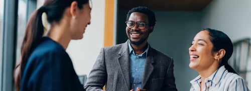 Trois personnes en tenue d'affaires se tiennent ensemble, souriant et conversant dans un cadre de bureau. L'atmosphère est conviviale et engageante.