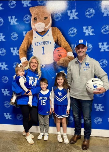 A family dressed in Kentucky Wildcats gear poses with a large mascot against a blue team-themed backdrop.